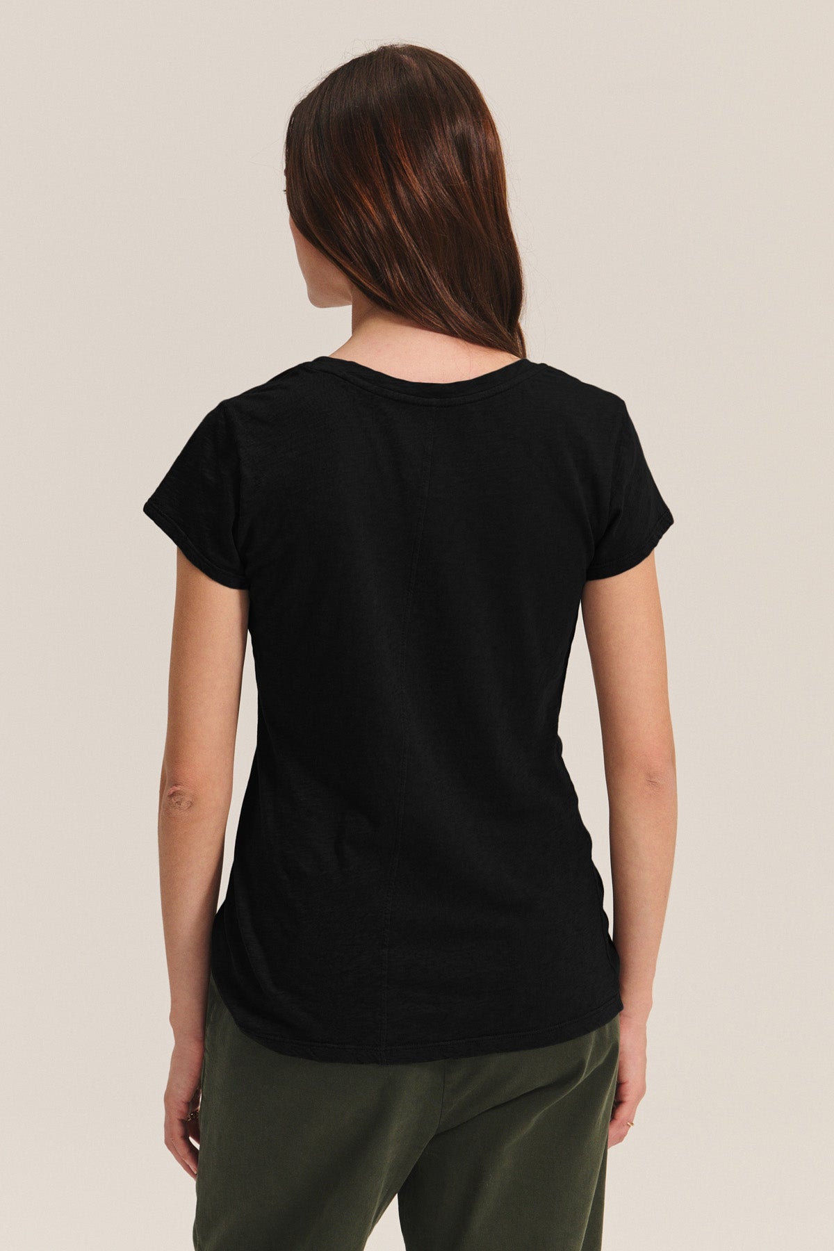 A person with their back to the camera wears the ODELIA TEE by Velvet by Graham & Spencer, a black short-sleeve t-shirt, paired with dark green pants against a plain background.-40292940710081