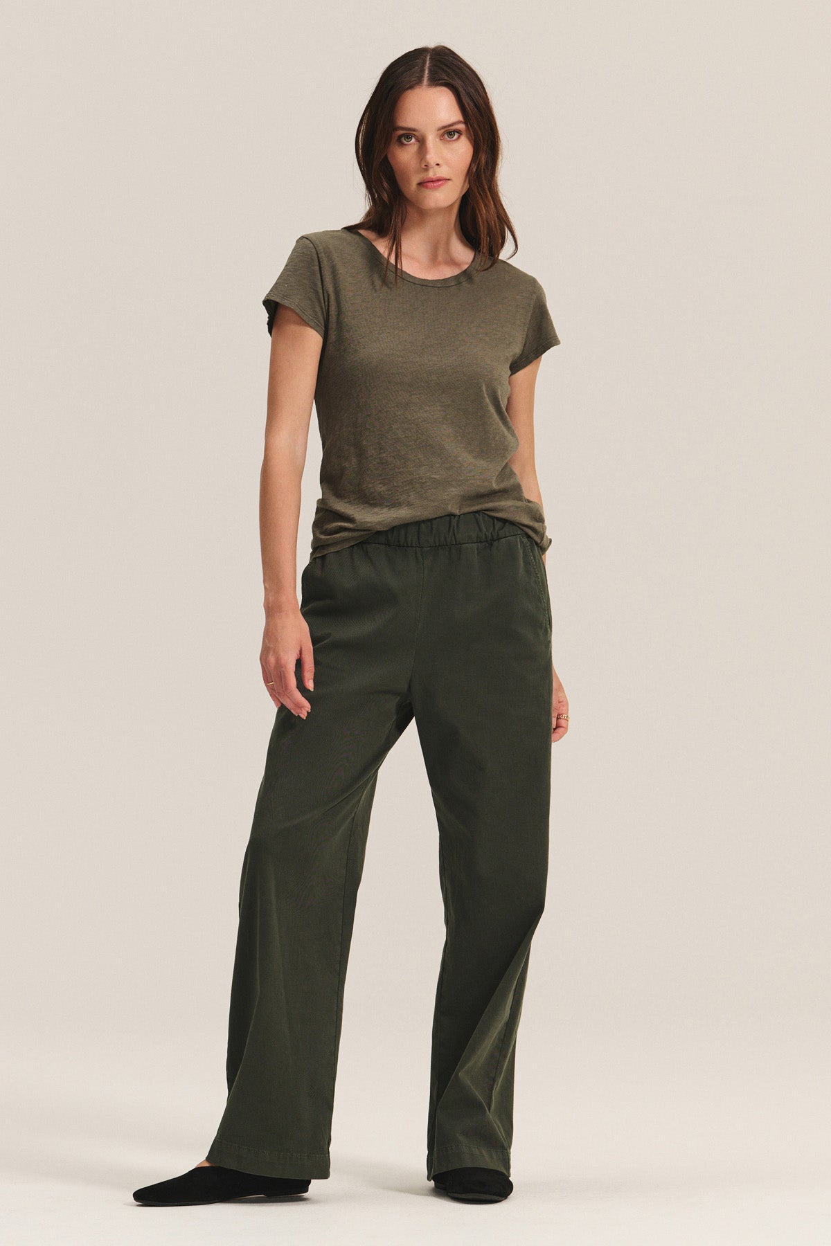A woman stands facing forward wearing the NAYA PANT by Velvet by Graham & Spencer—dark green wide-leg pants with an elastic waistband—paired with an olive green t-shirt and black shoes against a plain background.-40672443236545