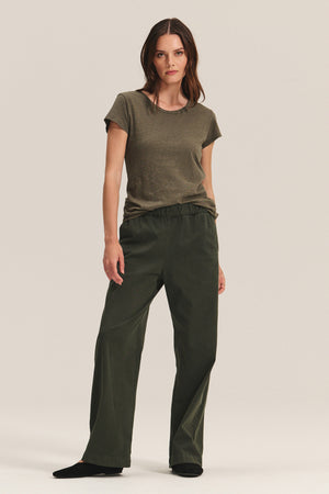 A woman stands against a neutral background wearing the ODELIA TEE by Velvet by Graham & Spencer in olive green, featuring a classic crew neckline, paired with dark green loose-fitting pants and black shoes.