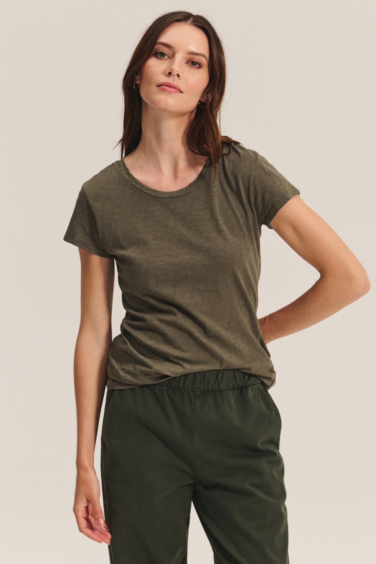 A woman stands against a plain light background, looking at the camera. She wears the ODELIA TEE by Velvet by Graham & Spencer in olive green with a classic crew neckline, paired with dark green pants.-40598548906177