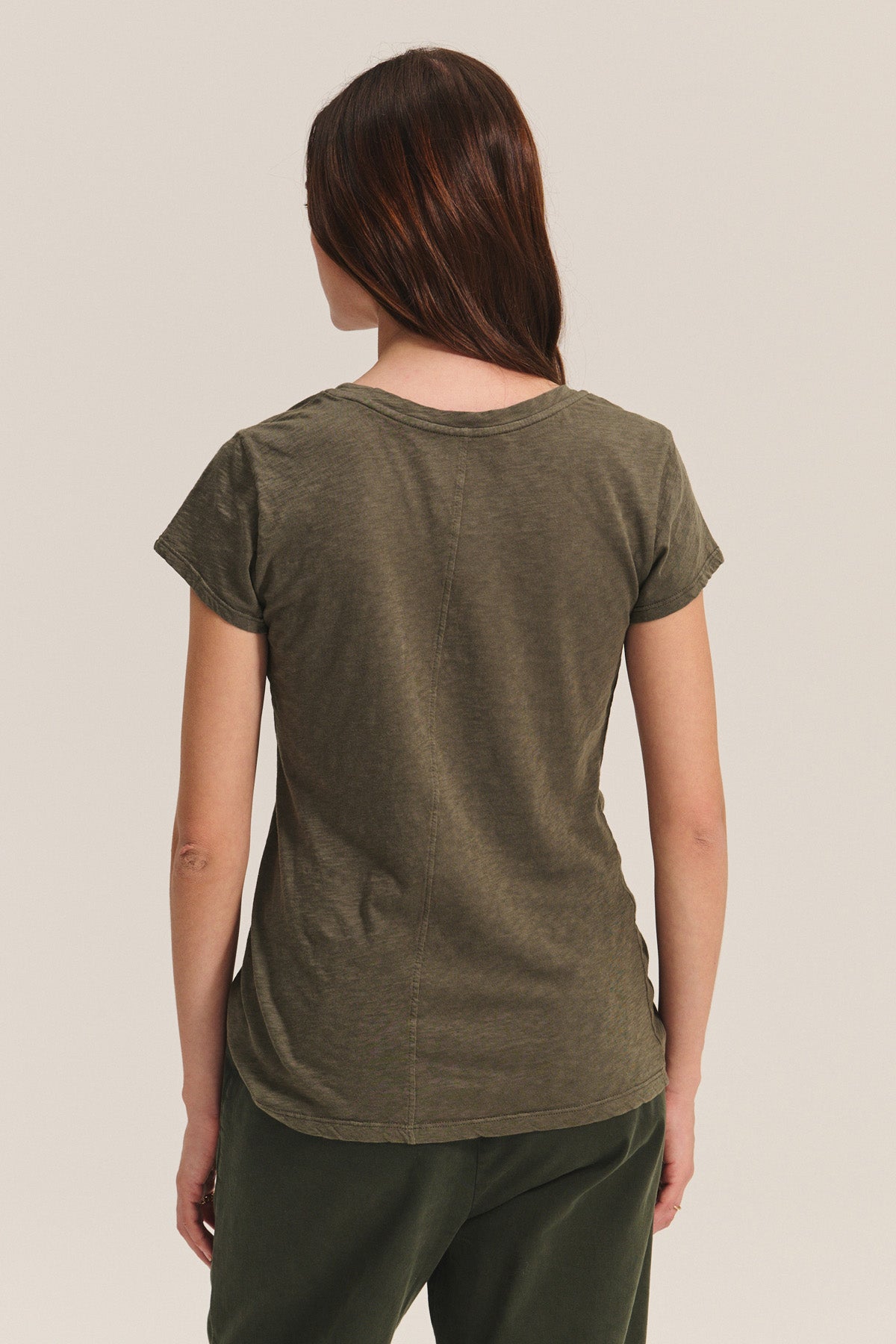 A woman with brown hair, wearing the ODELIA TEE by Velvet by Graham & Spencer in olive green and dark pants, stands facing away from the camera against a plain background.