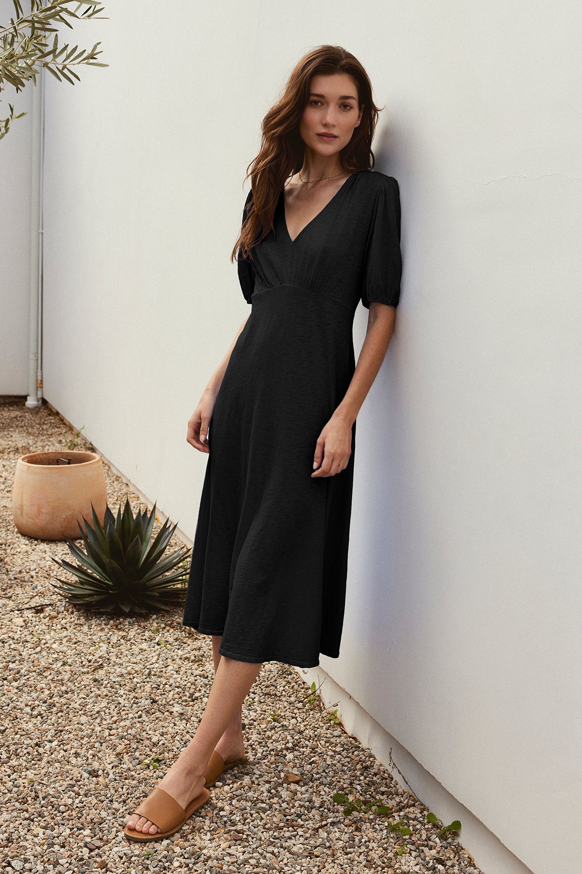 A woman in a figure-skimming black PARKER COTTON SLUB MIDI DRESS by Velvet by Graham & Spencer with puffed sleeves and tan sandals stands against a white wall, next to a small garden with plants in pots.-37185421967553