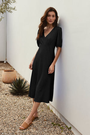 A woman in a figure-skimming black PARKER COTTON SLUB MIDI DRESS by Velvet by Graham & Spencer with puffed sleeves and tan sandals stands against a white wall, next to a small garden with plants in pots.
