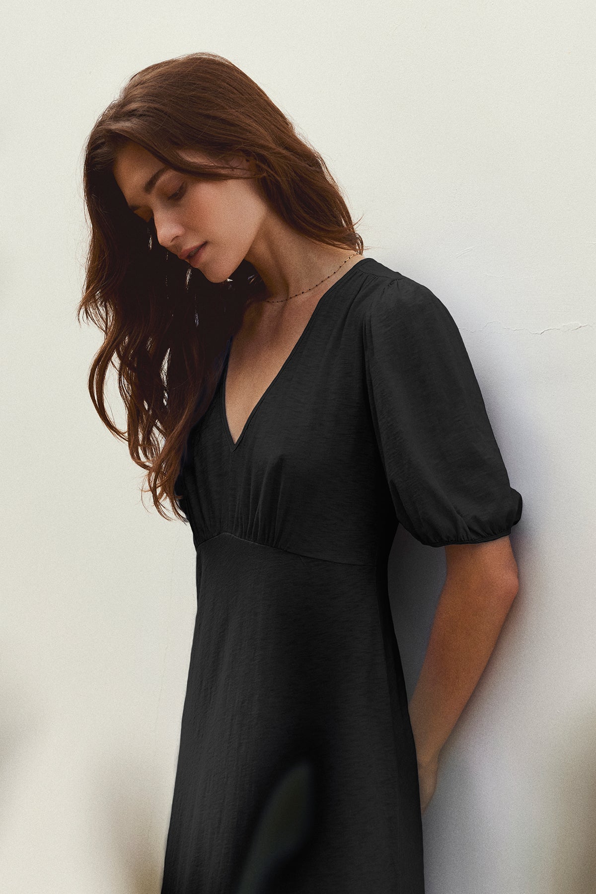 A woman with long brown hair wearing a black PARKER COTTON SLUB MIDI DRESS by Velvet by Graham & Spencer stands against a plain white wall, looking downwards.-37185422000321
