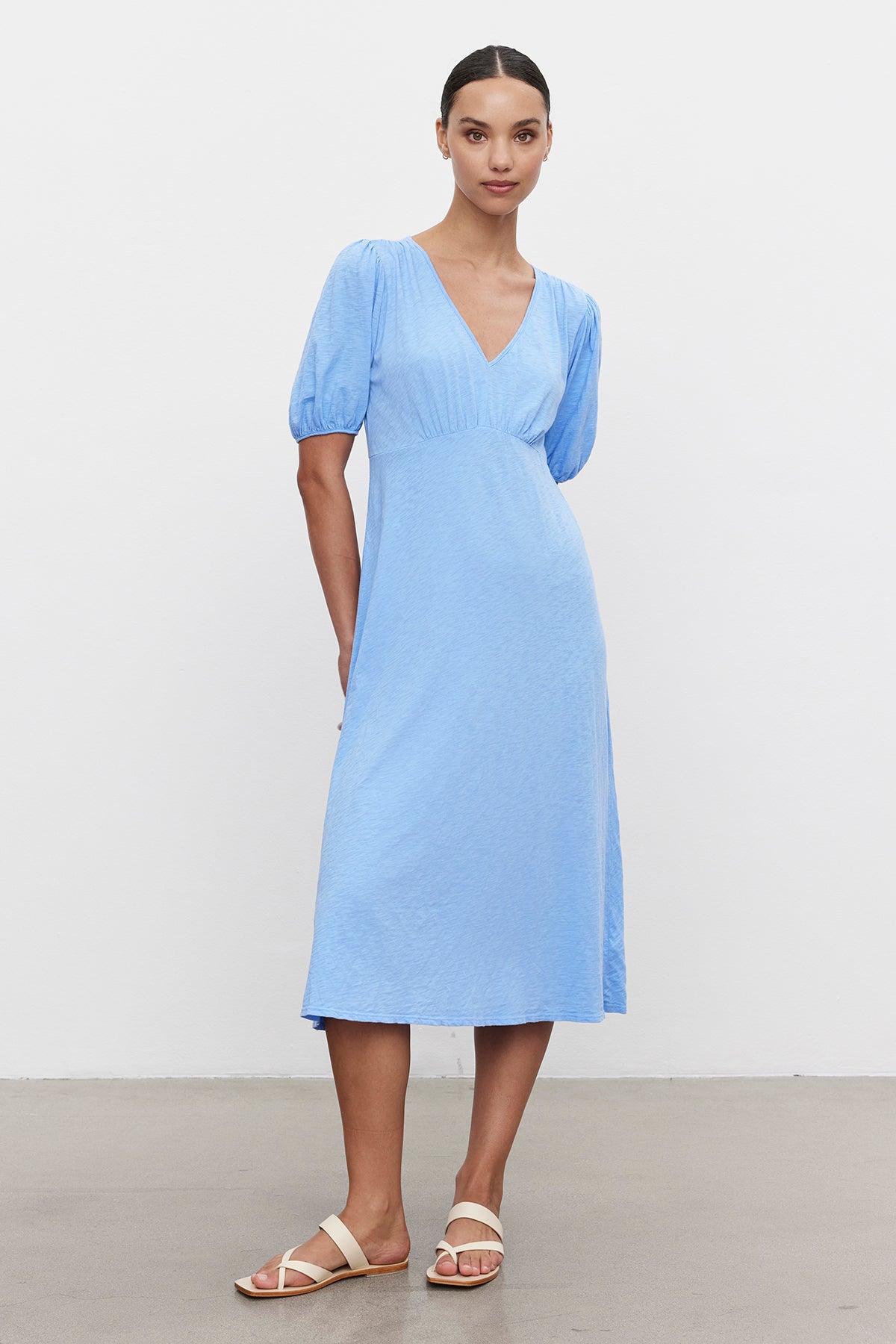 A woman in a Velvet by Graham & Spencer PARKER COTTON SLUB MIDI DRESS with puff sleeves and a v-neckline, paired with white sandals, stands against a white background.-36691474743489
