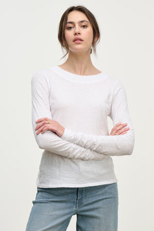 A woman with long brown hair in a low bun models the PRIYA TEE by Velvet by Graham & Spencer—a plain white long-sleeve shirt—paired with light blue jeans, standing arms crossed against a light background.