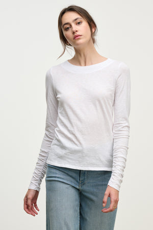 A woman wears the versatile PRIYA TEE by Velvet by Graham & Spencer—a relaxed fit, plain white long-sleeve shirt—with light blue jeans, standing against a neutral background.