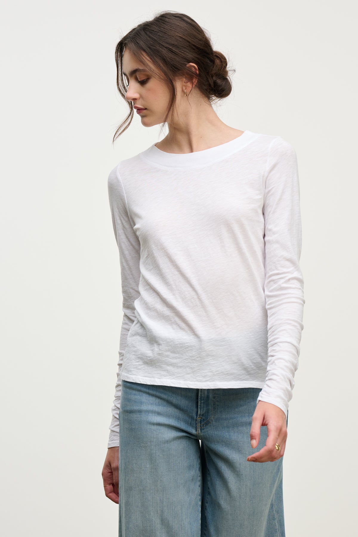 A woman models the PRIYA TEE by Velvet by Graham & Spencer—a relaxed fit, long-sleeved white top—paired with light blue jeans, standing slightly angled against a plain background to showcase this versatile wardrobe staple.-39814912770241