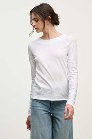 A woman models the PRIYA TEE by Velvet by Graham & Spencer—a relaxed fit, long-sleeved white top—paired with light blue jeans, standing slightly angled against a plain background to showcase this versatile wardrobe staple.