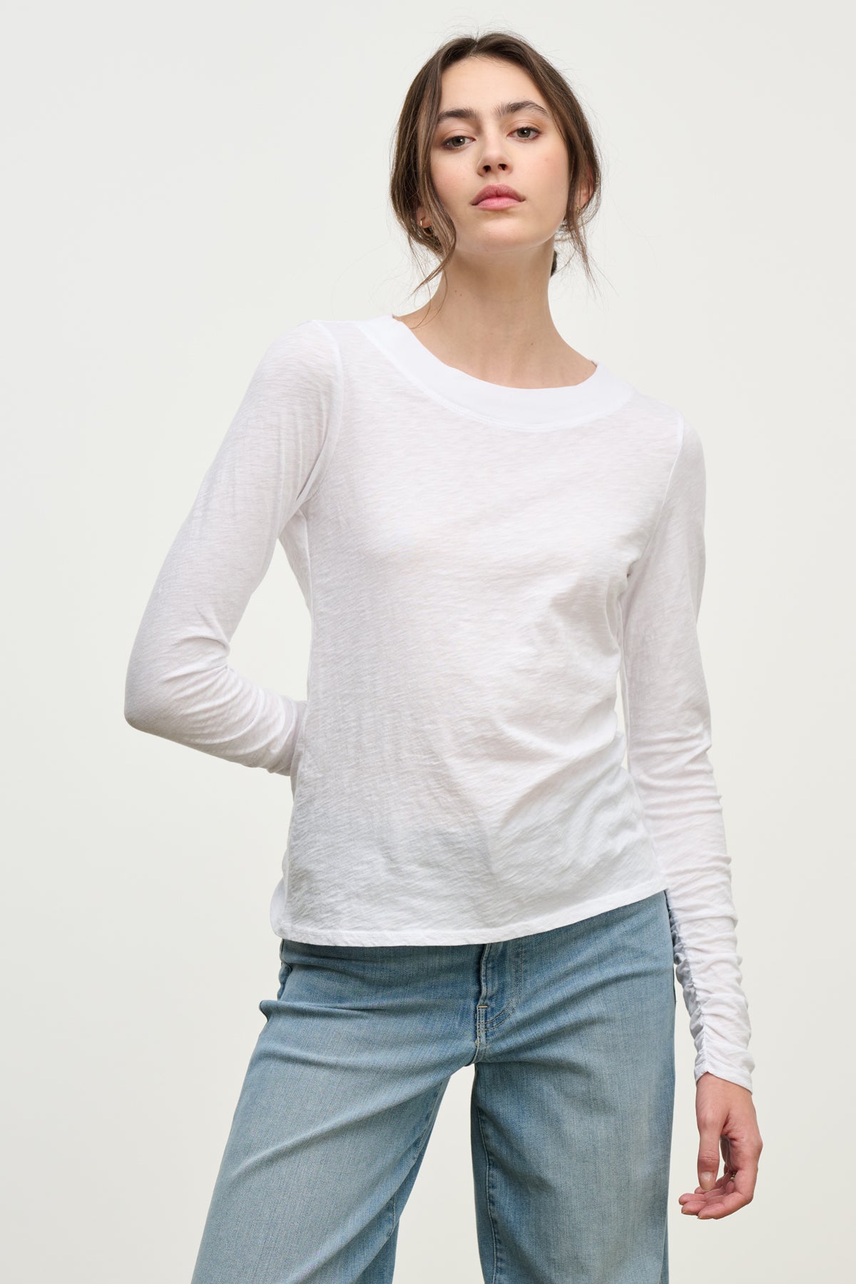 A woman wearing the Velvet by Graham & Spencer PRIYA TEE, a relaxed fit plain white long-sleeve shirt, with light blue jeans stands against a neutral background.-39814912737473