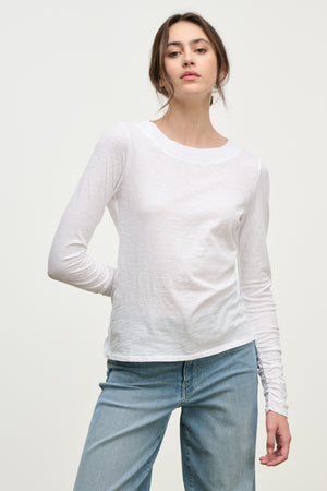 A woman wearing the Velvet by Graham & Spencer PRIYA TEE, a relaxed fit plain white long-sleeve shirt, with light blue jeans stands against a neutral background.