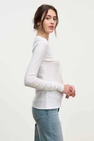 A woman stands in profile against a plain background, wearing the PRIYA TEE by Velvet by Graham & Spencer—a white, relaxed fit long-sleeve shirt—paired with blue jeans.
