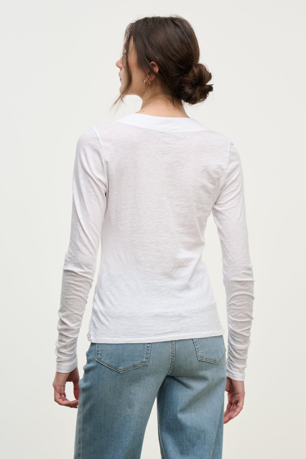 A woman with brown hair in a low bun wears the PRIYA TEE by Velvet by Graham & Spencer—a relaxed fit, long-sleeve white shirt—paired with light blue jeans, standing with her back to the camera against a plain background.-39814912671937