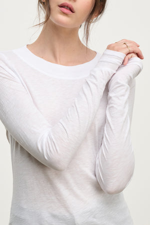 A woman models the PRIYA TEE by Velvet by Graham & Spencer—a sheer white, relaxed-fit long-sleeve shirt—arms slightly crossed, minimal jewelry, against a neutral background; an effortlessly versatile wardrobe staple.