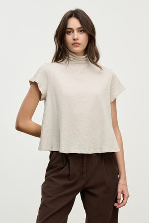 A woman stands against a plain background, wearing the SAVI TEE by Velvet by Graham & Spencer—a beige relaxed turtleneck mock neck top—paired with loose brown pants, highlighting a boxy silhouette.