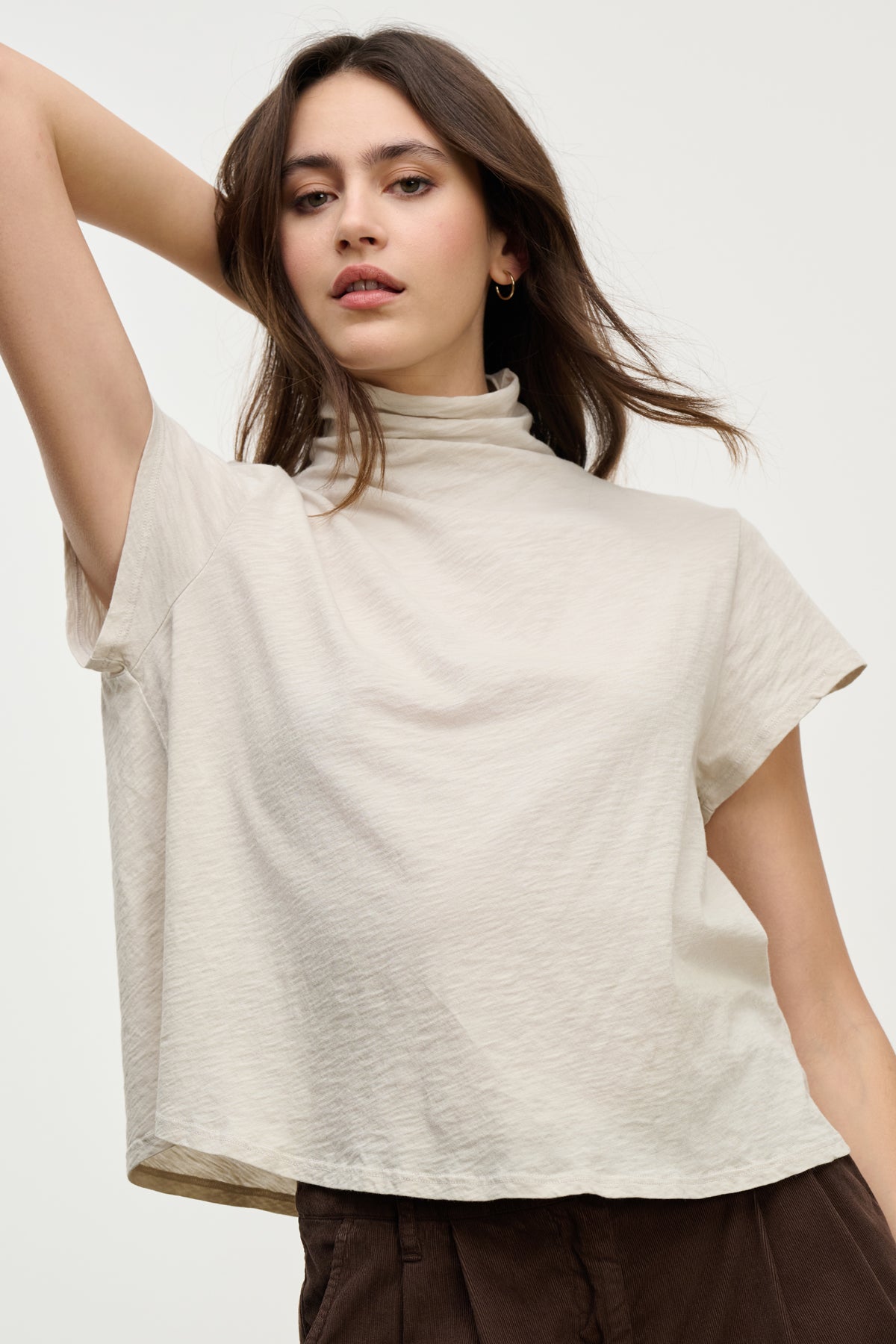 A woman with long brown hair models the SAVI TEE by Velvet by Graham & Spencer, a relaxed turtleneck cotton slub tee in light beige, paired with dark brown pants, posing with one arm raised against a plain light background.-39800374001857