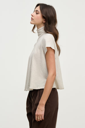A woman stands in profile against a plain background, wearing the SAVI TEE by Velvet by Graham & Spencer—a relaxed, short-sleeve turtleneck with a boxy silhouette—paired with dark pants.