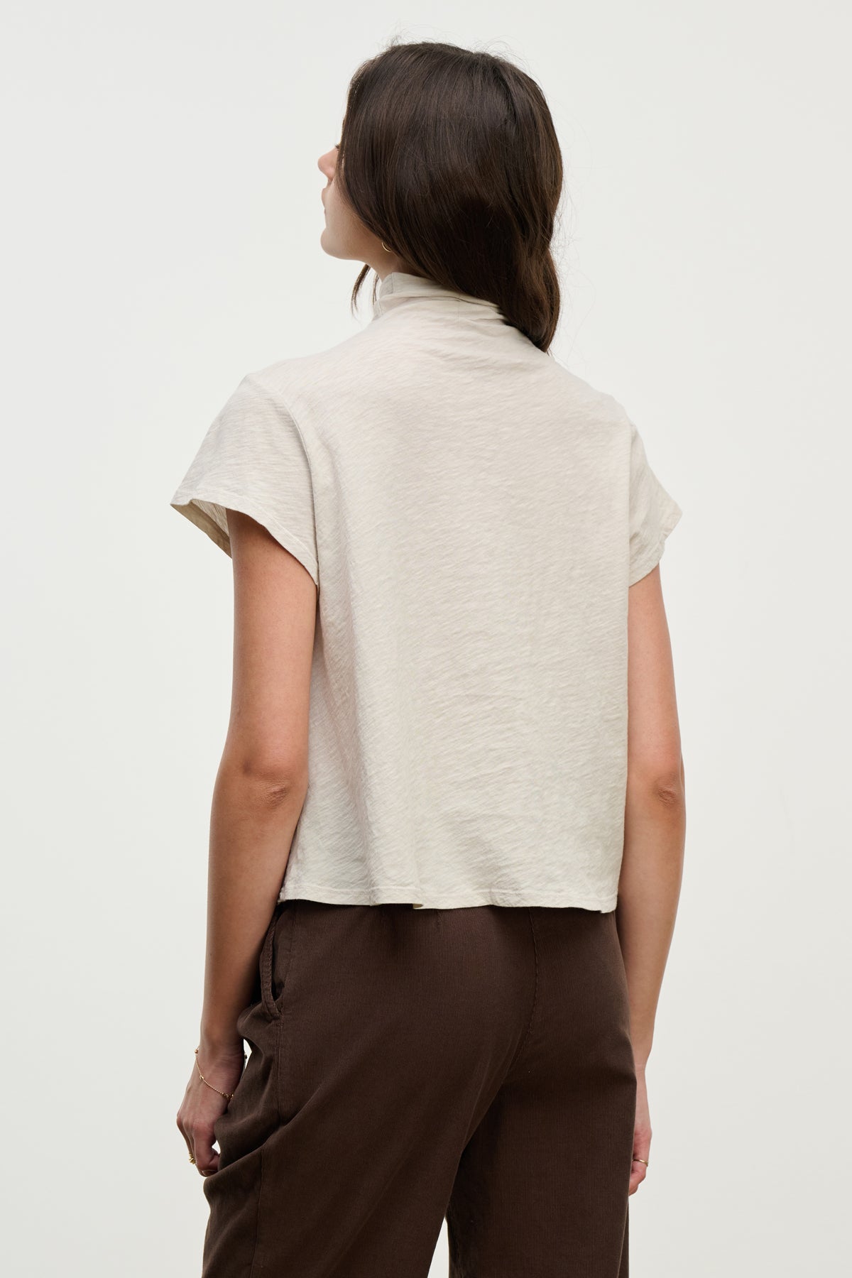 A person stands facing away, wearing the SAVI TEE by Velvet by Graham & Spencer—a light beige cotton slub tee with a boxy silhouette—paired with dark brown pants against a plain light background.-39800373936321