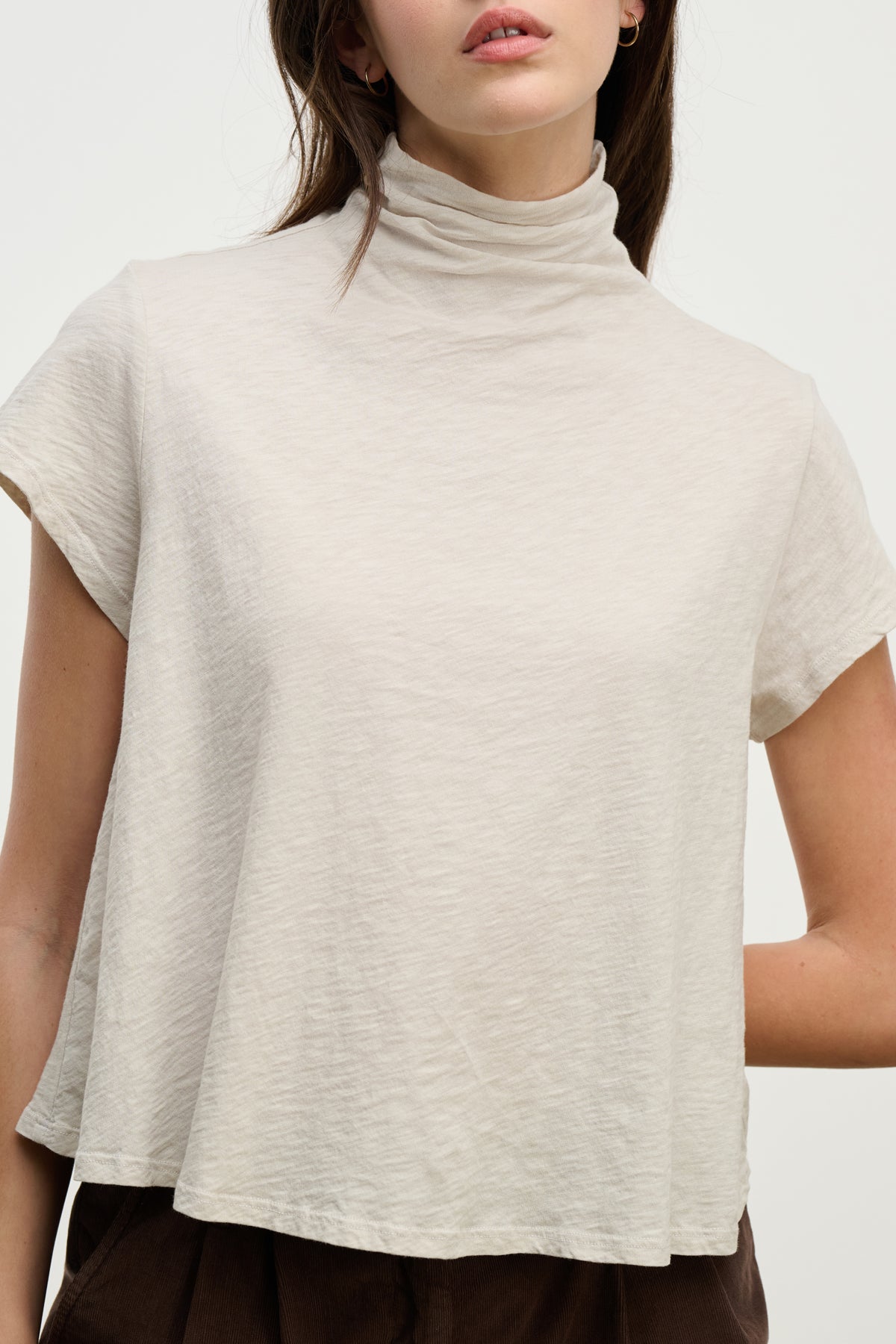 A person wearing the SAVI TEE by Velvet by Graham & Spencer, a relaxed light beige short-sleeve turtleneck with a boxy silhouette, paired with dark brown pants; shown from shoulders to waist.-39800373903553