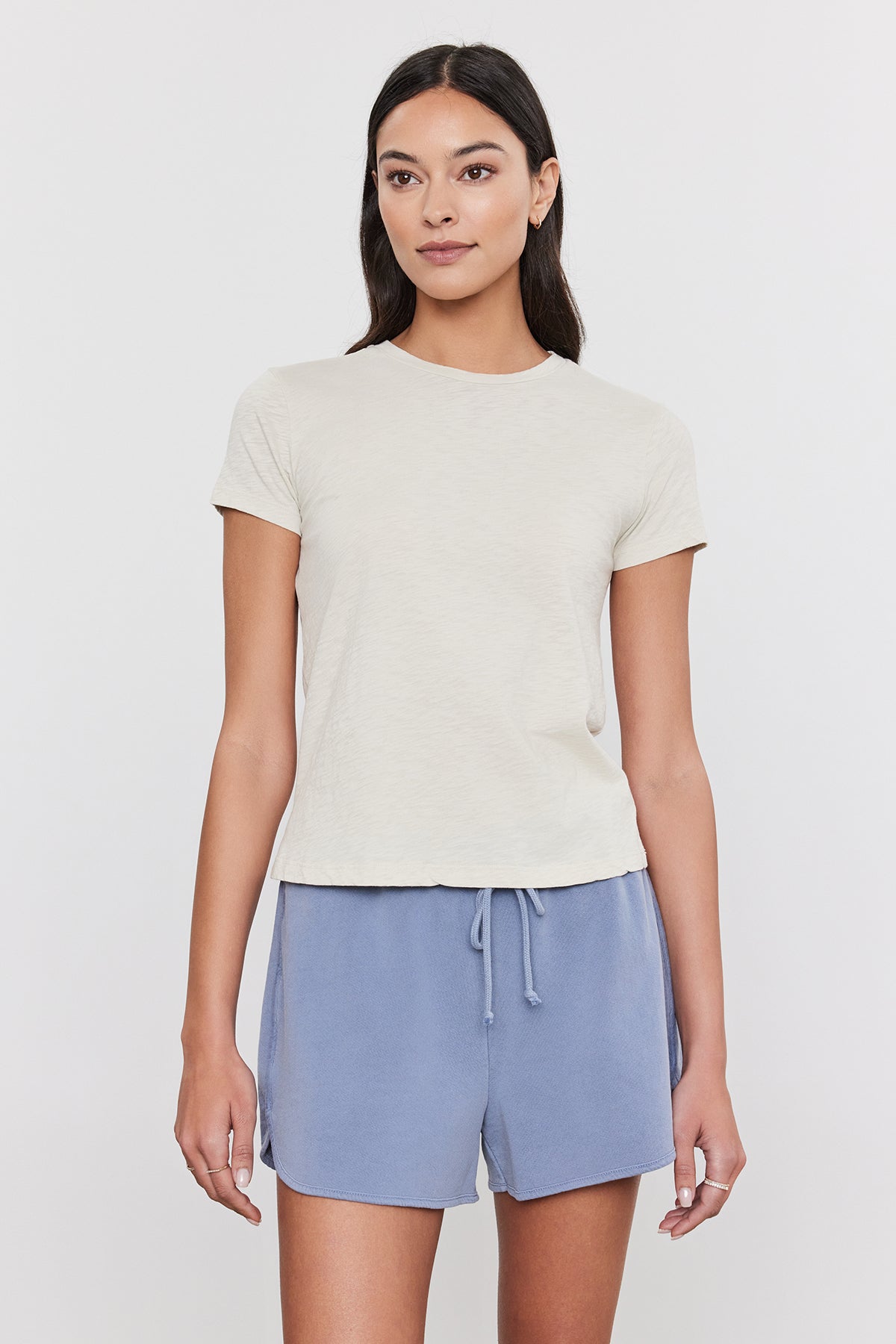   A person with long dark hair is wearing a light beige SIERRA CREW NECK TEE by Velvet by Graham & Spencer and light blue drawstring shorts, standing against a plain white background, exuding a subtle retro vibe. 