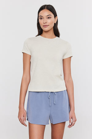 A person with long dark hair is wearing a light beige SIERRA CREW NECK TEE by Velvet by Graham & Spencer and light blue drawstring shorts, standing against a plain white background, exuding a subtle retro vibe.