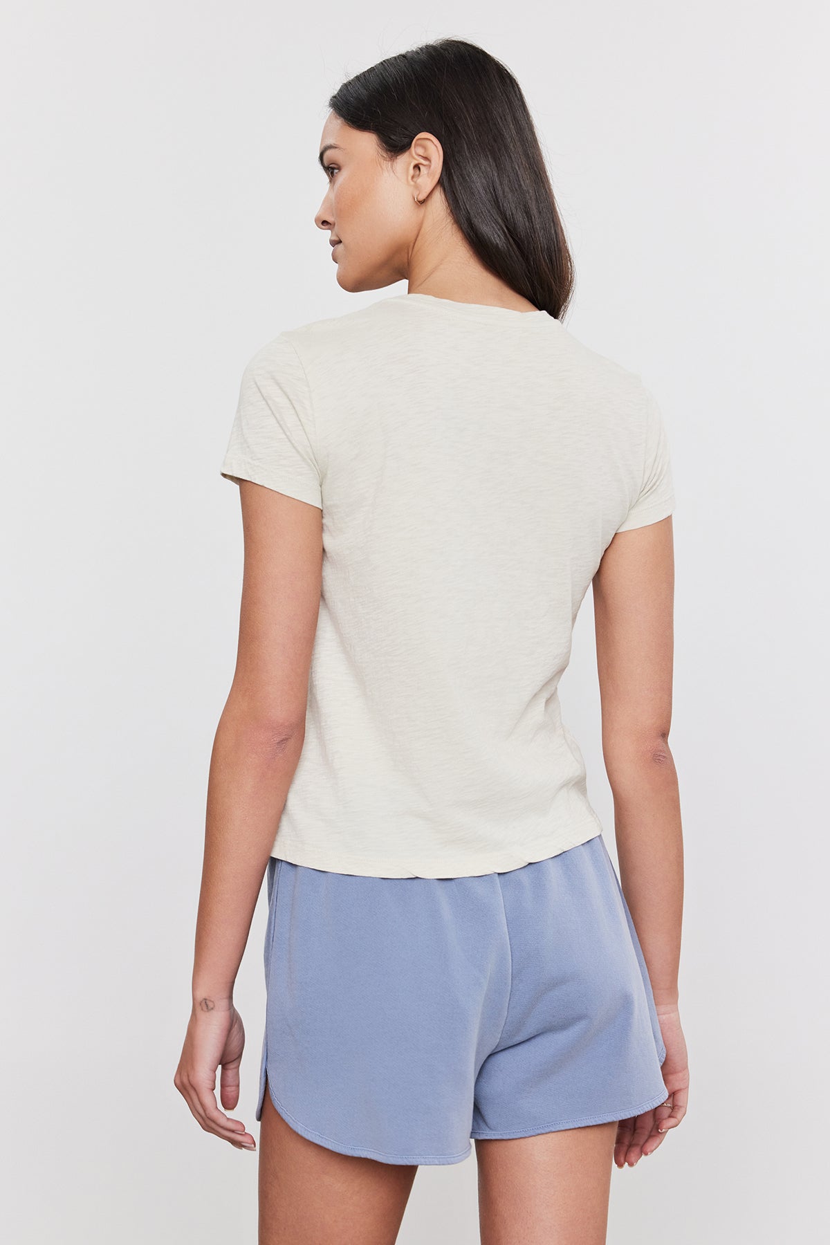   A woman with long dark hair stands with her back to the camera, wearing a short-sleeve beige SIERRA CREW NECK TEE by Velvet by Graham & Spencer and light blue shorts against a plain background, capturing a subtle retro vibe. 