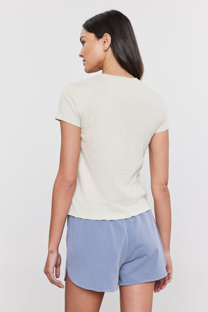 A woman with long dark hair stands with her back to the camera, wearing a short-sleeve beige SIERRA CREW NECK TEE by Velvet by Graham & Spencer and light blue shorts against a plain background, capturing a subtle retro vibe.