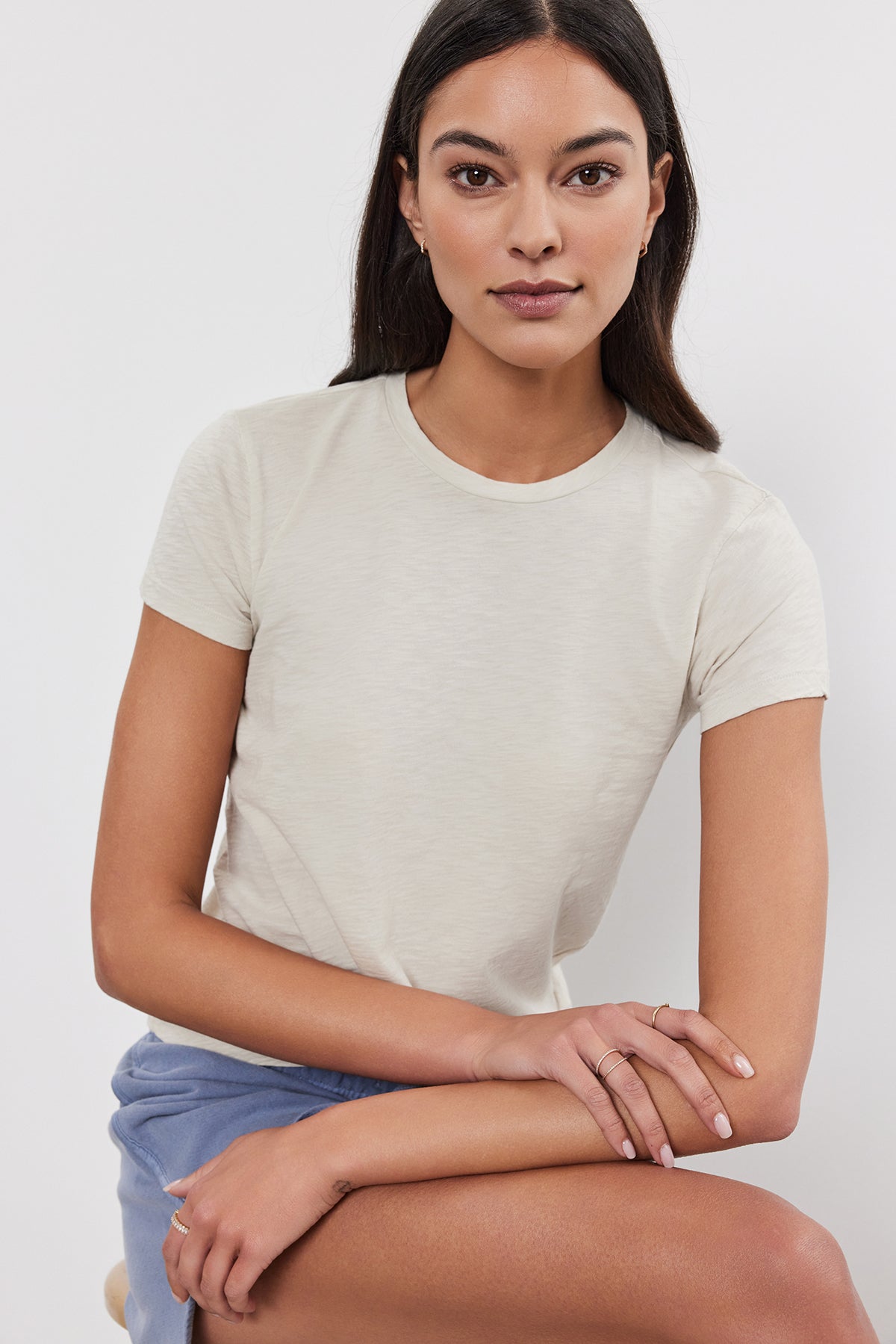   A person with long dark hair, wearing a short-sleeved beige SIERRA CREW NECK TEE by Velvet by Graham & Spencer and light blue shorts, sits on a stool against a plain background. 