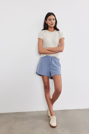 A person standing against a white wall, wearing a white SIERRA CREW NECK TEE from Velvet by Graham & Spencer, blue shorts, and light-colored shoes. They are with their arms crossed and leaning slightly back, exuding a retro vibe.