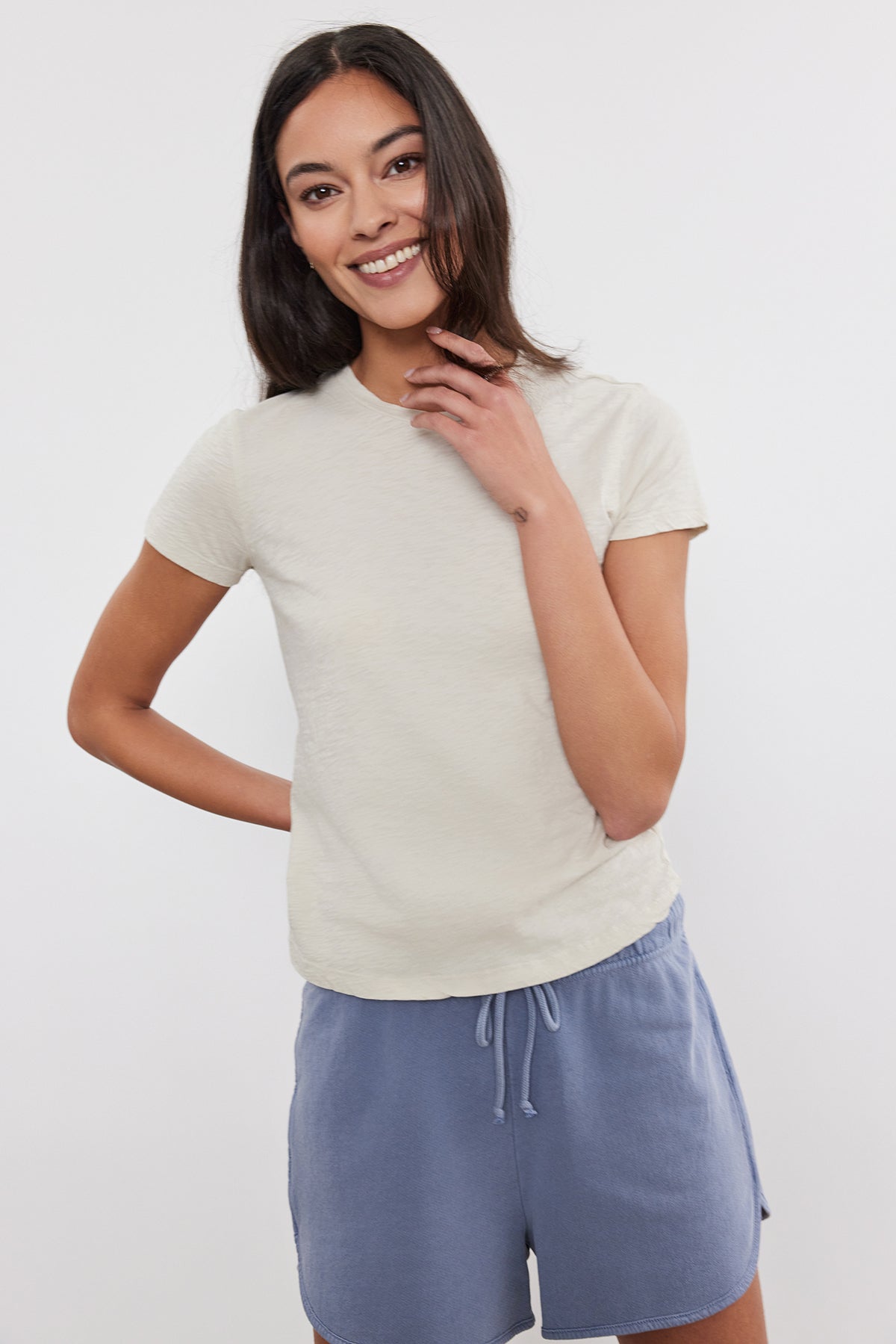   A woman with long dark hair, wearing a beige SIERRA CREW NECK TEE by Velvet by Graham & Spencer and blue drawstring shorts, smiles and poses with one hand on her hip and the other touching her face. 