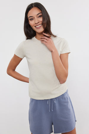 A woman with long dark hair, wearing a beige SIERRA CREW NECK TEE by Velvet by Graham & Spencer and blue drawstring shorts, smiles and poses with one hand on her hip and the other touching her face.