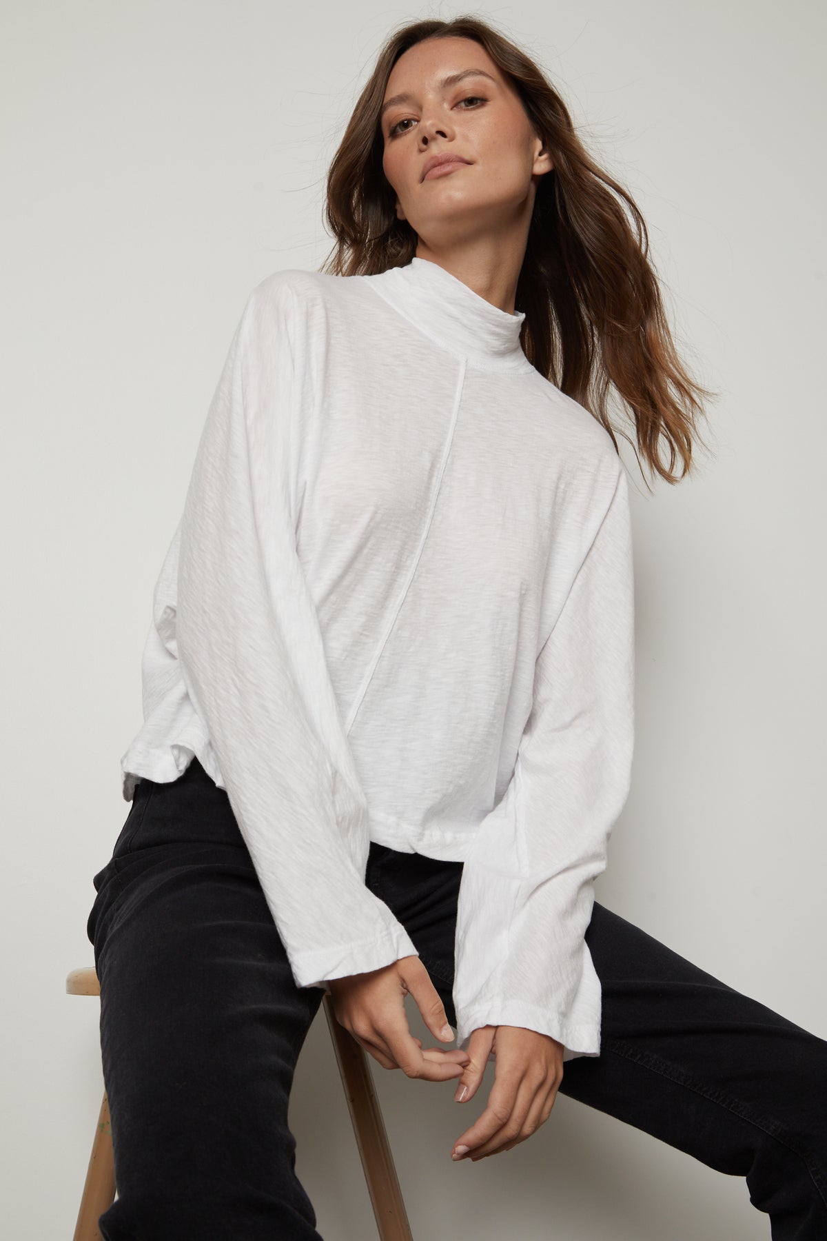 A person with long hair wears a white STACEY MOCK NECK TEE by Velvet by Graham & Spencer and black pants, seated against a plain background.-36328480768193