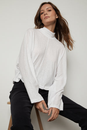 A person with long hair wears a white STACEY MOCK NECK TEE by Velvet by Graham & Spencer and black pants, seated against a plain background.