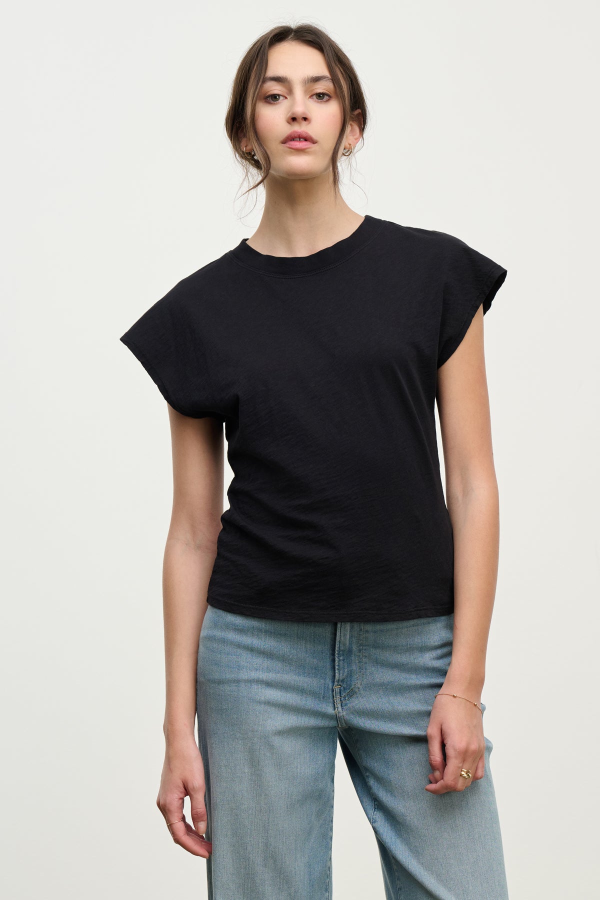A woman faces forward against a plain background, wearing the Velvet by Graham & Spencer STERLING TEE—a black cotton slub top with dolman short sleeves—paired with light blue jeans and a neutral expression.-39800415649985