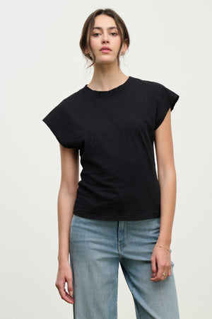 A woman faces forward against a plain background, wearing the Velvet by Graham & Spencer STERLING TEE—a black cotton slub top with dolman short sleeves—paired with light blue jeans and a neutral expression.