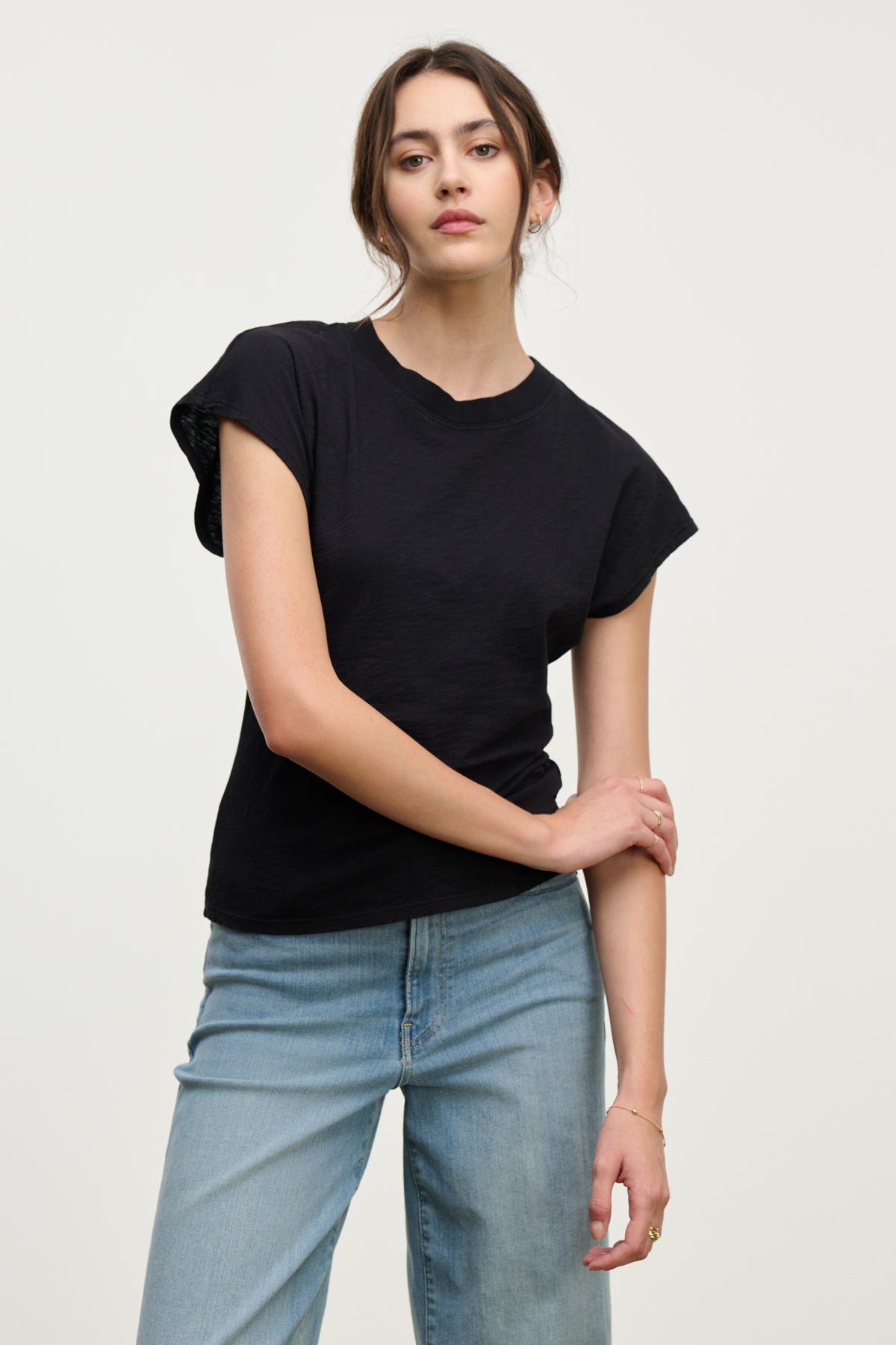 A woman stands against a plain background wearing the STERLING TEE by Velvet by Graham & Spencer, a black cotton slub dolman short-sleeve top, paired with light blue jeans; one arm bent, her hand on her opposite forearm.