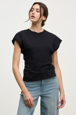 A person wearing the STERLING TEE by Velvet by Graham & Spencer, a black cotton slub top with dolman short sleeves, and light blue jeans stands against a plain light background.