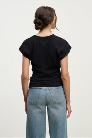A woman with brown hair in a loose bun, wearing the Velvet by Graham & Spencer STERLING TEE in black and blue jeans, stands facing away against a plain white background.