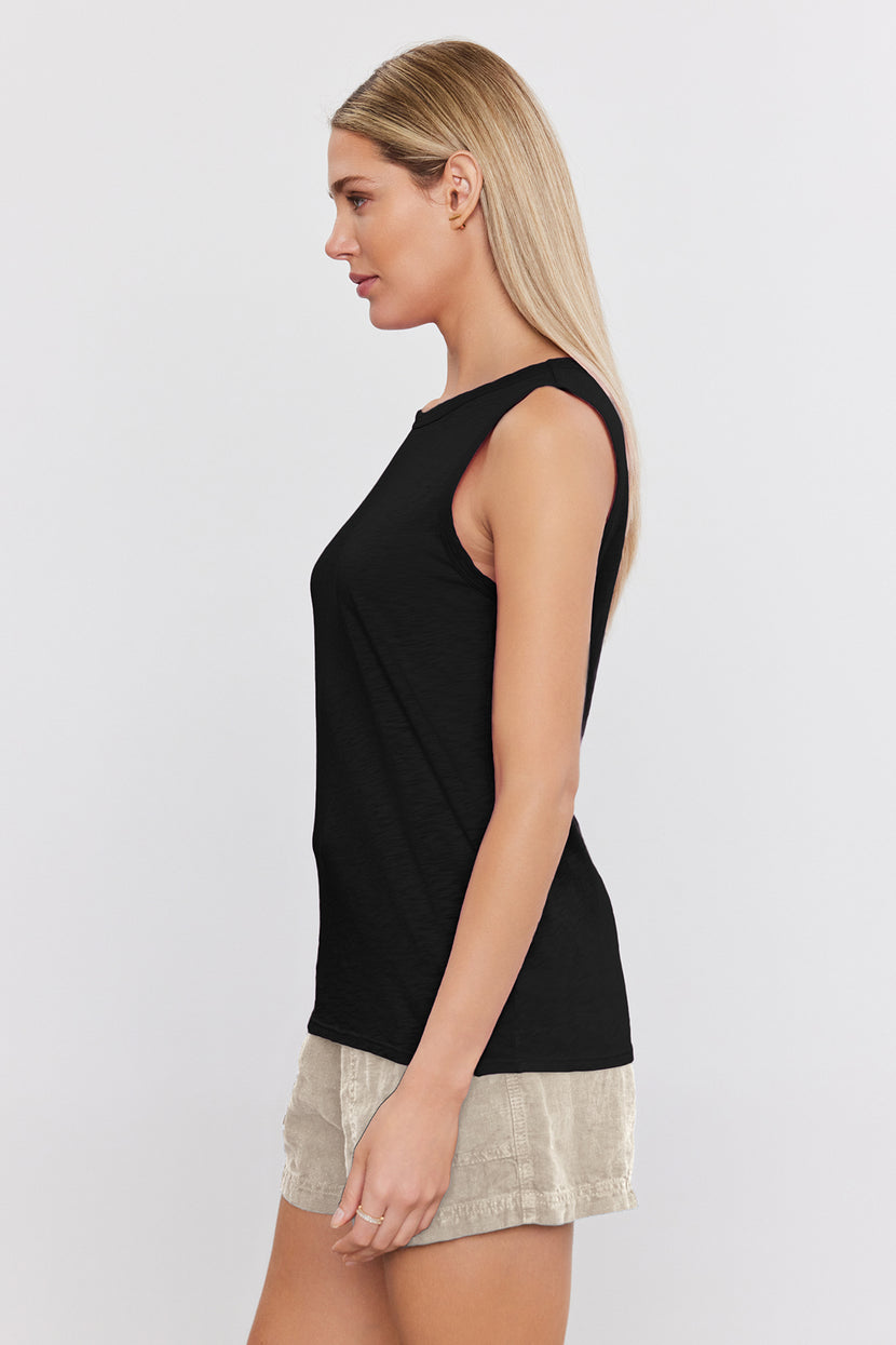 A woman with long blonde hair is shown in profile, wearing a sleeveless black top and light-colored shorts. The TAURUS TANK TOP by Velvet by Graham & Spencer and her textured cotton slub shorts make for a versatile staple outfit. She stands against a plain white background.