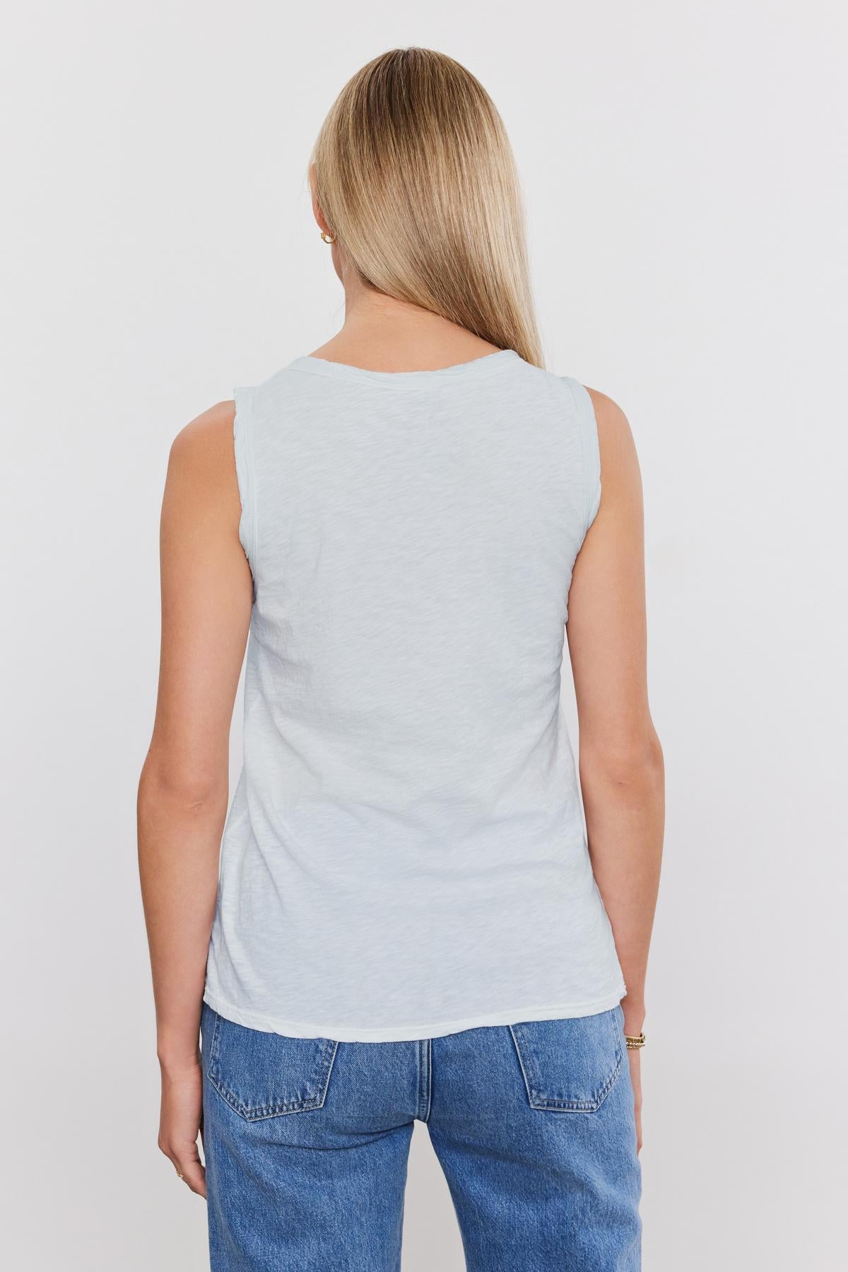   A person with long blond hair wears the TAURUS TANK TOP by Velvet by Graham & Spencer—a light gray, textured cotton slub sleeveless top—and blue jeans, viewed from the back against a plain white background. 