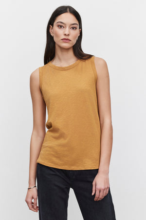 A person with long dark hair wears the TAURUS TANK TOP by Velvet by Graham & Spencer, a sleeveless, mustard-colored, crew neck top, paired with black pants while standing against a plain white background.
