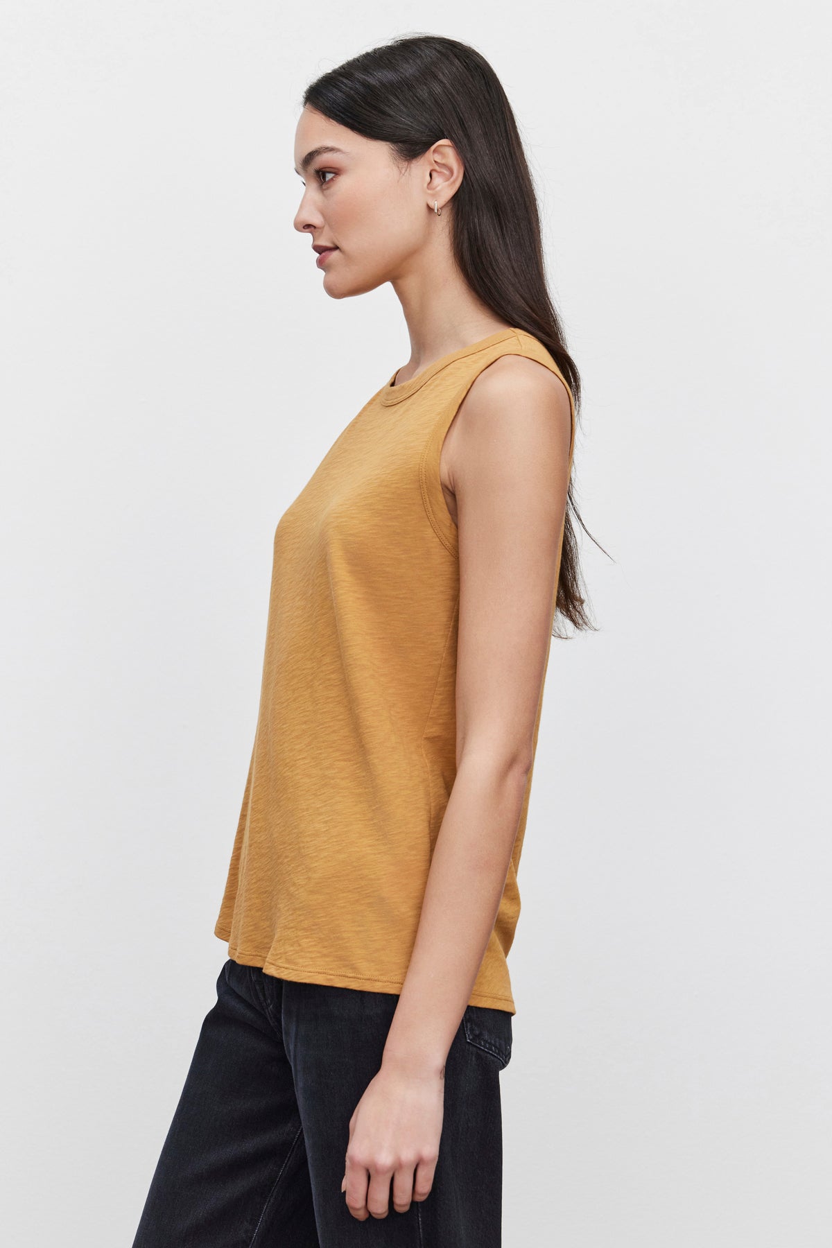 A woman with long, dark hair stands in profile, wearing the sleeveless TAURUS TANK TOP from Velvet by Graham & Spencer, which is made from cotton slub and comes in a mustard color. She pairs it with dark pants against a plain white background. The top's fabric and design make it an essential piece for any wardrobe.-37629572710593