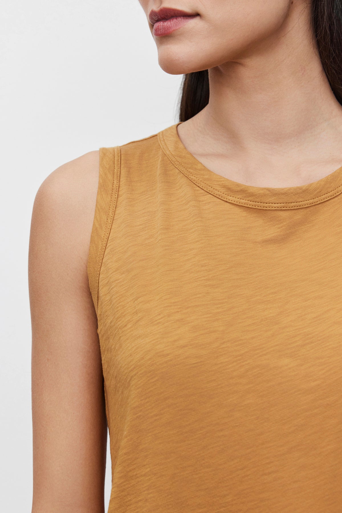   Close-up of a person wearing the TAURUS TANK TOP in mustard-yellow from Velvet by Graham & Spencer. The crew neck adds a touch of simplicity to this essential piece. The person has long dark hair, and their face is partially visible. 