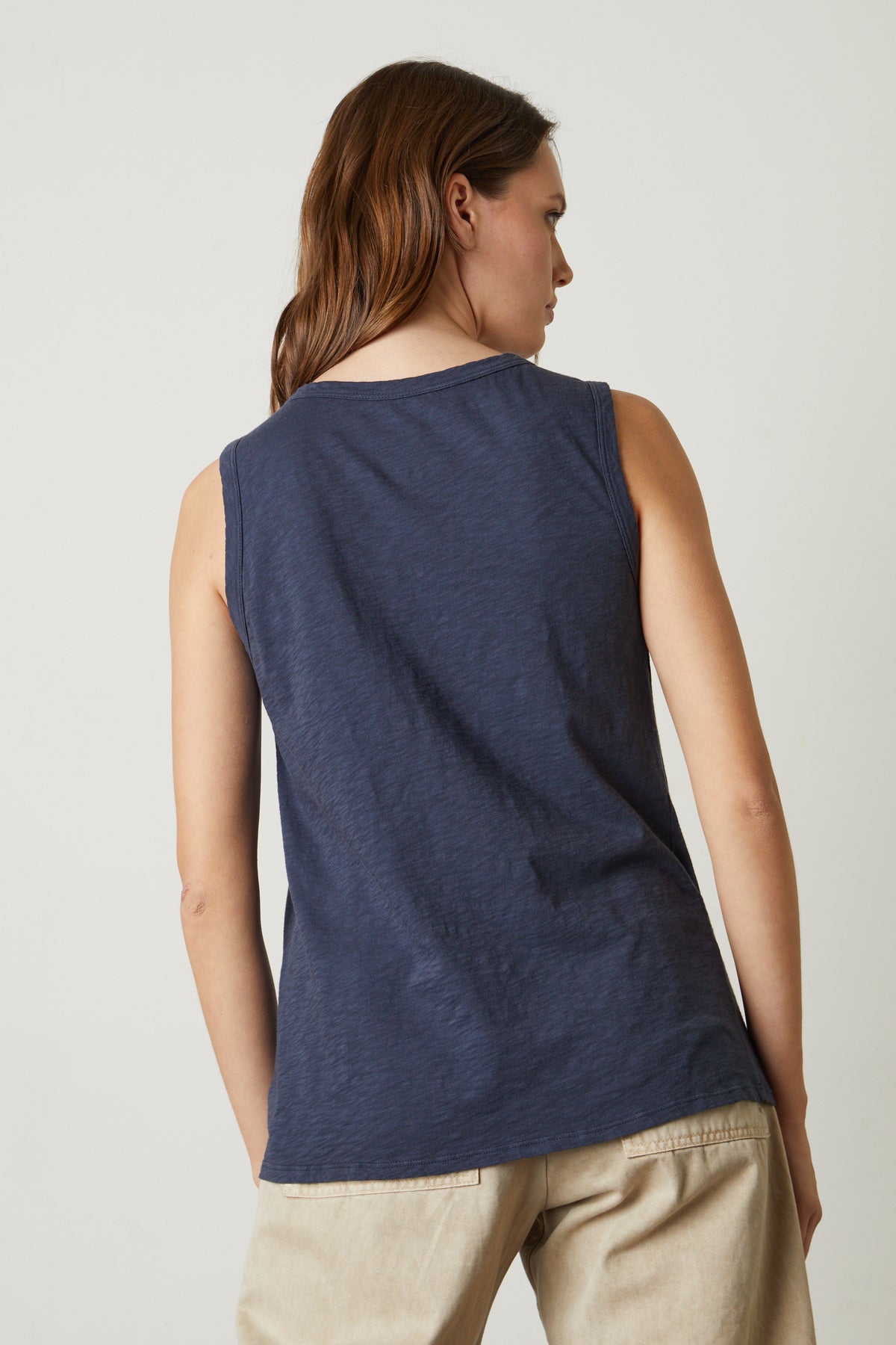   A person with brown hair stands facing away, wearing a timeless crew neck navy blue TAURUS TANK TOP from Velvet by Graham & Spencer made of textured cotton slub and beige pants against a plain background. 