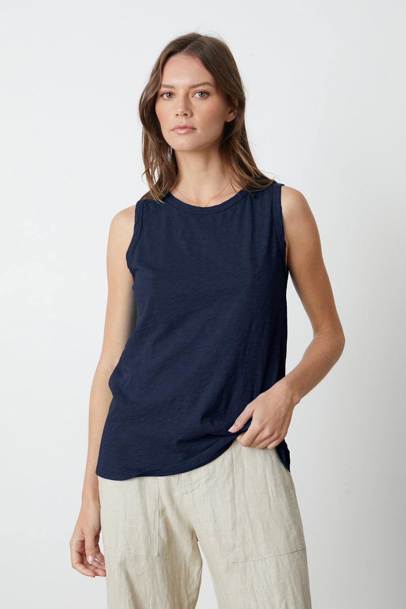 A woman with shoulder-length brown hair is wearing a sleeveless navy blue top and beige pants. The timeless crew neck and textured cotton slub TAURUS TANK TOP by Velvet by Graham & Spencer makes her outfit a versatile staple as she stands against a plain white background.