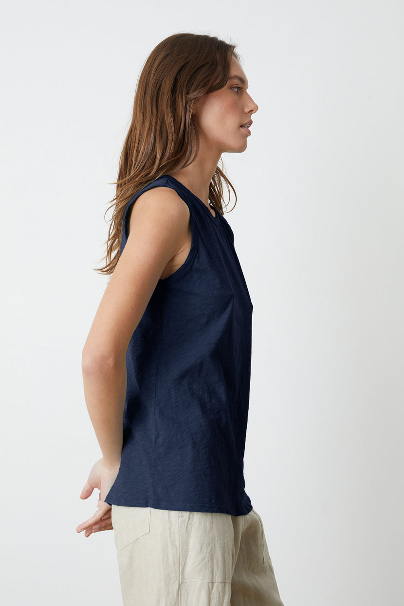 A woman with long brown hair is standing in profile, wearing a sleeveless navy TAURUS TANK TOP by Velvet by Graham & Spencer made of textured cotton slub and light-colored pants. Her arms are positioned behind her back against a plain white background.