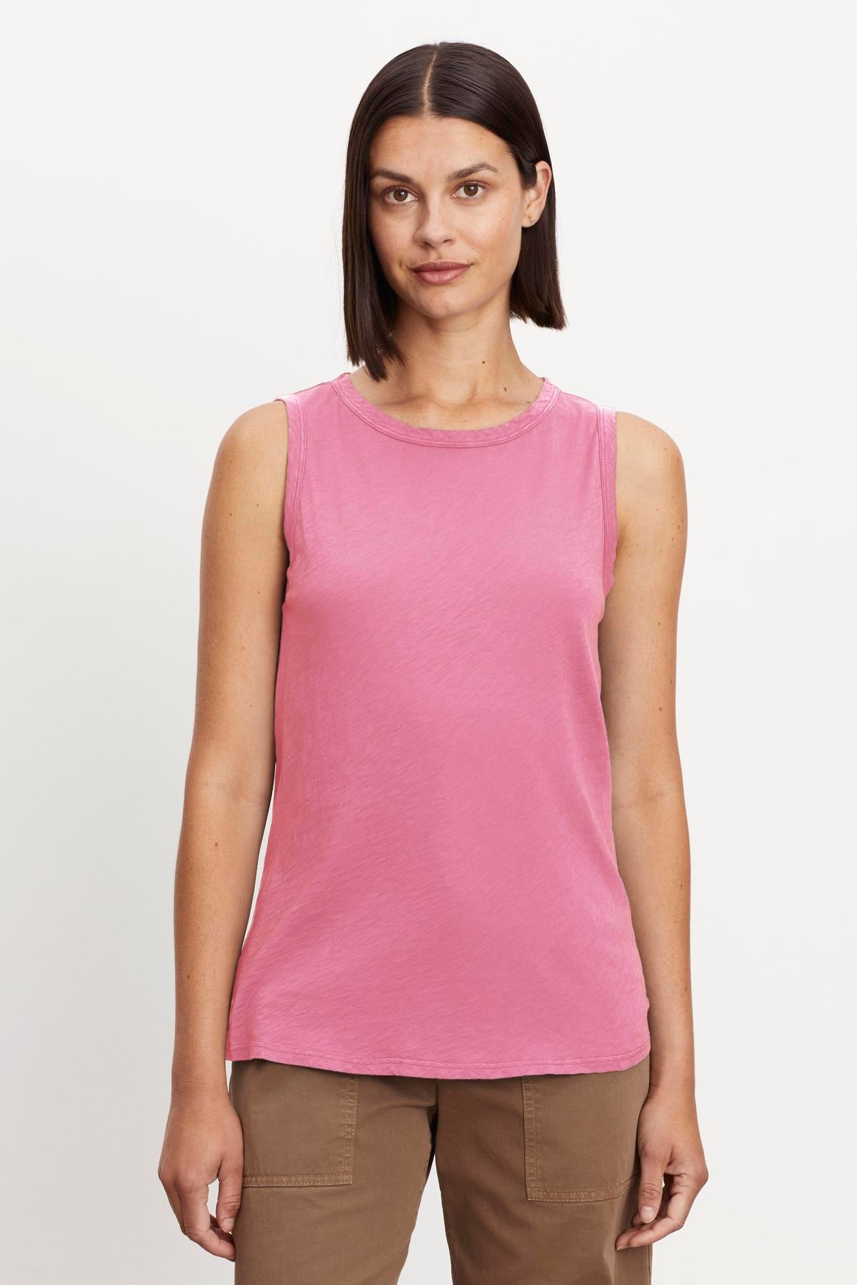 A woman with shoulder-length brown hair wearing a sleeveless pink TAURUS TANK TOP by Velvet by Graham & Spencer and brown pants stands against a plain background.-37249949466817