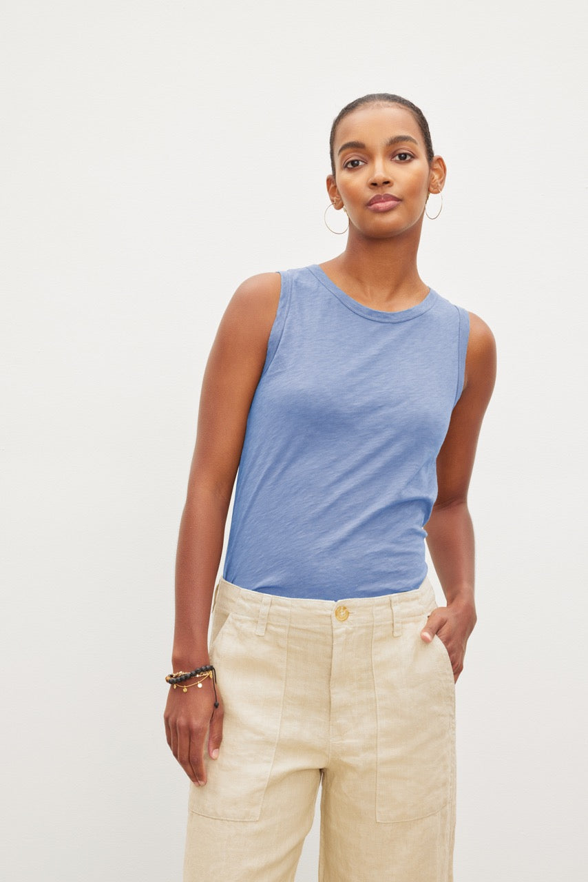   A woman in a blue crew-neck TAURUS COTTON SLUB TANK and beige trousers standing against a white background, looking directly at the camera. 
