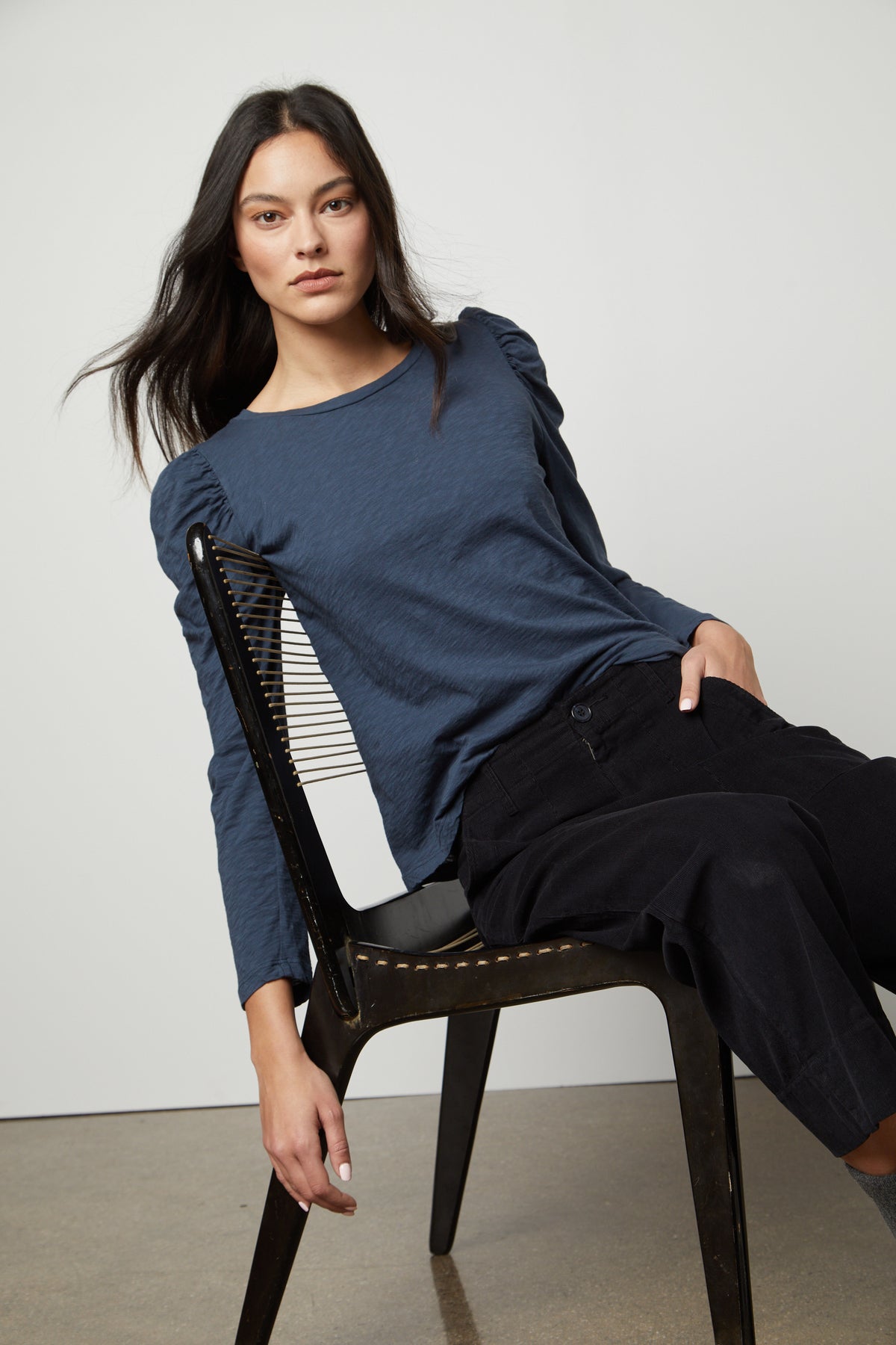 A woman is sitting on a chair wearing a TORA CREW NECK TEE by Velvet by Graham & Spencer, blue top and black pants.-26872376721601