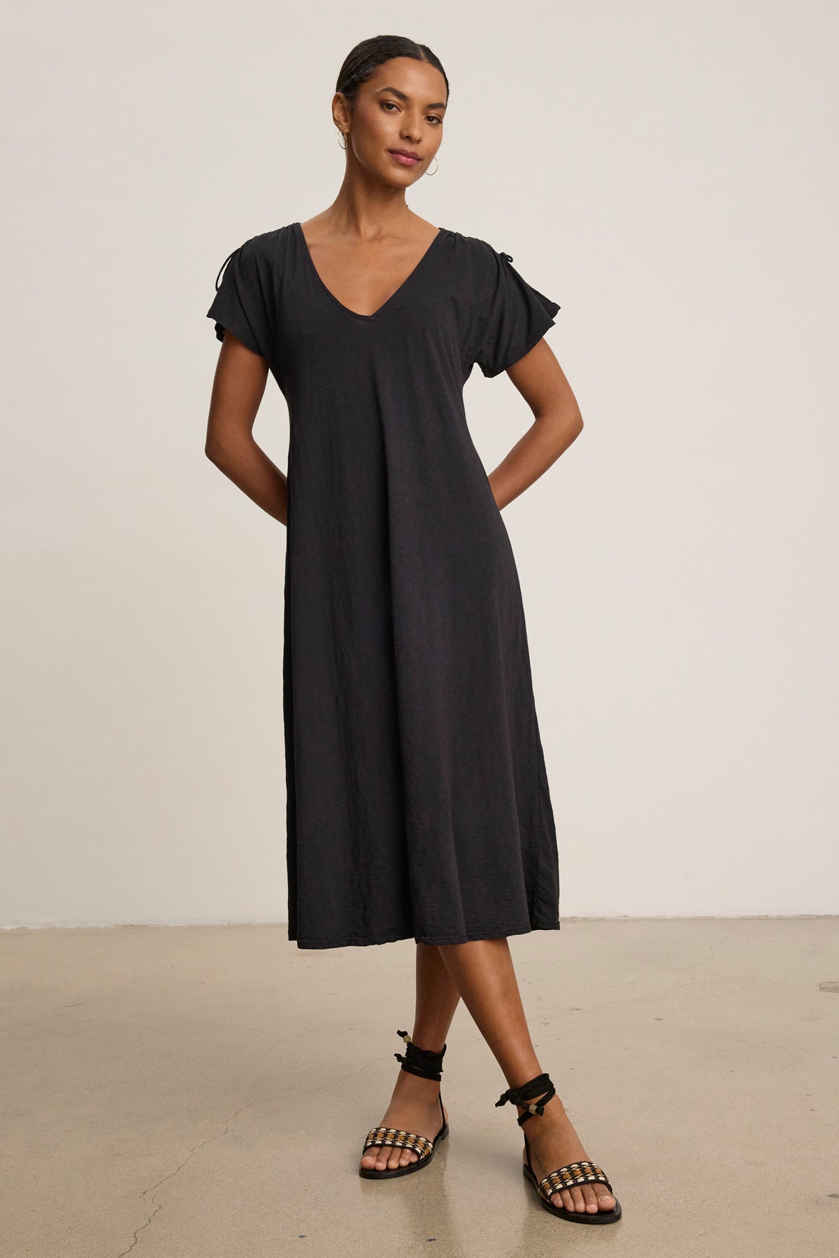   A woman stands against a plain background wearing the VERONICA DRESS by Velvet by Graham & Spencer—a loose-fitting, short-sleeved black t-shirt dress with a v-neckline—paired with black strappy sandals. 